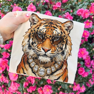 K&ouml;nnte beinhalten: Ein cremefarbenes quadratisches Kissen mit einer detaillierten Illustration eines Tigers mit Leopardenmuster-Sonnenbrille und einer Diamantkette. Der Tiger hat orange und schwarze Streifen. Der Hintergrund ist eine lebendige Darstellung von rosa Blumen.