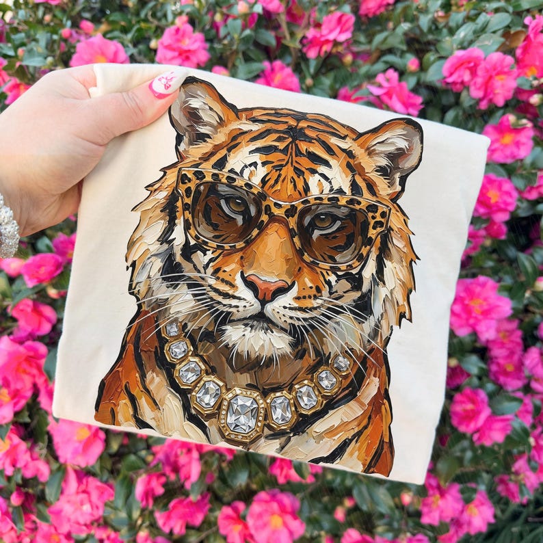 K&ouml;nnte beinhalten: Ein cremefarbenes quadratisches Kissen mit einer detaillierten Illustration eines Tigers mit Leopardenmuster-Sonnenbrille und einer Diamantkette. Der Tiger hat orange und schwarze Streifen. Der Hintergrund ist eine lebendige Darstellung von rosa Blumen.