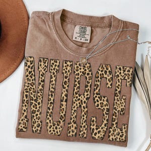 May include: A brown Comfort Colors t-shirt with the word "NURSE" in large leopard print letters. A silver necklace with a pendant rests on the shirt. The shirt is folded and displayed on a white surface.