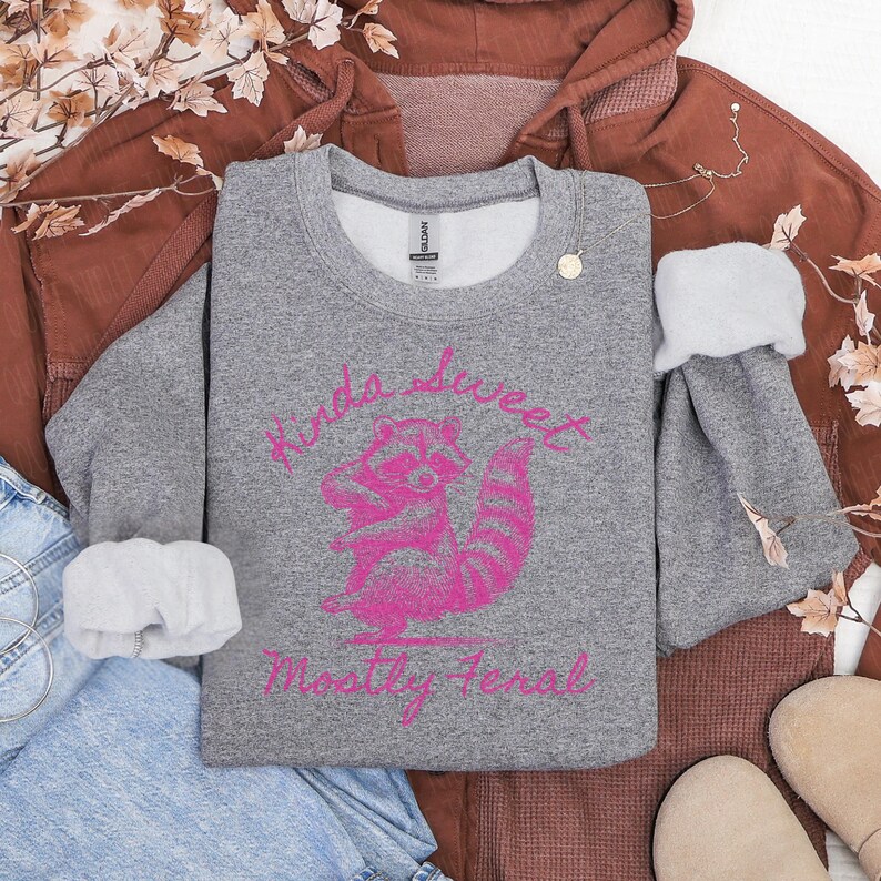 May include: A gray sweatshirt with the words "Kinda Sweet, Mostly Feral" in pink script, along with a pink raccoon graphic. The sweatshirt is folded on top of a brown hoodie and blue jeans.