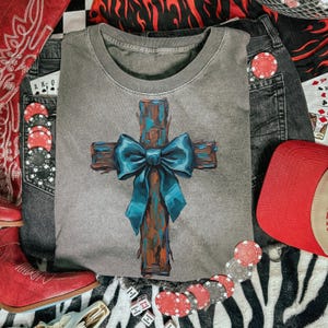 Peut inclure: Un t-shirt gris &agrave; col rond avec une croix peinte et un n&oelig;ud bleu sarcelle. L'image comprend une casquette rouge, un jean, des jetons de poker, des d&eacute;s et des bottes de cowboy rouges, sur un fond z&eacute;br&eacute;.