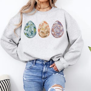 Peut inclure: Un sweat-shirt gris clair avec trois &oelig;ufs de P&acirc;ques peints. Les &oelig;ufs sont bleus, beiges et violets, avec des coups de pinceau textur&eacute;s. Le sweat-shirt a des manches longues et un col rond.