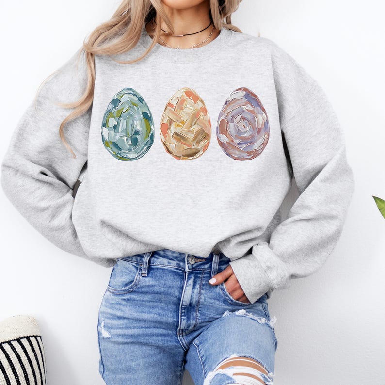Peut inclure: Un sweat-shirt gris clair avec trois &oelig;ufs de P&acirc;ques peints. Les &oelig;ufs sont bleus, beiges et violets, avec des coups de pinceau textur&eacute;s. Le sweat-shirt a des manches longues et un col rond.