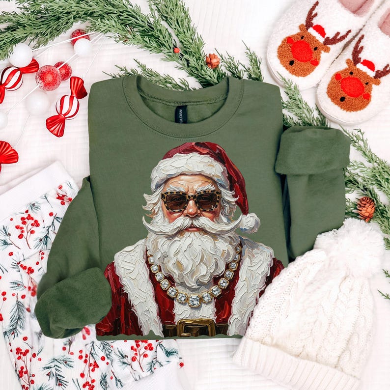 May include: An olive green sweatshirt featuring a graphic print of Santa Claus wearing leopard print sunglasses and a thick gold chain. The Santa illustration has a textured, painterly style with a white beard, red hat, and red coat.