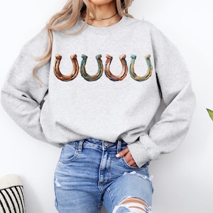 May include: A light gray sweatshirt featuring four horseshoe graphics in shades of brown, green, and orange. The horseshoe design is centered on the chest. The model is wearing blue jeans.