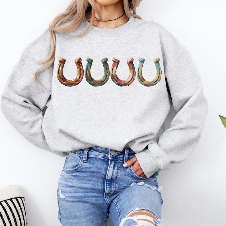 May include: A light gray sweatshirt featuring four horseshoe graphics in shades of brown, green, and orange. The horseshoe design is centered on the chest. The model is wearing blue jeans.