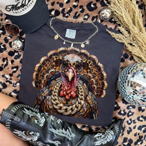 May include: A dark gray t-shirt featuring a graphic of a turkey wearing leopard print sunglasses and a thick gold chain. The turkey has detailed brown and tan feathers. The shirt is displayed with a black baseball cap, sunglasses, and a gold necklace.
