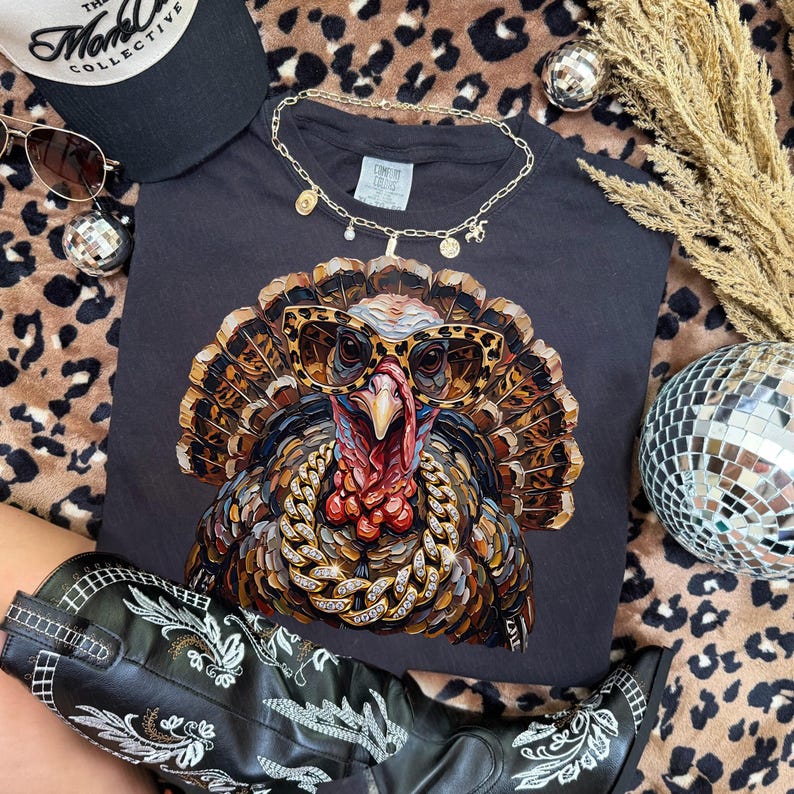 May include: A dark gray t-shirt featuring a graphic of a turkey wearing leopard print sunglasses and a thick gold chain. The turkey has detailed brown and tan feathers. The shirt is displayed with a black baseball cap, sunglasses, and a gold necklace.