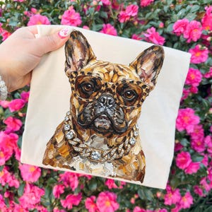 May include: A cream-colored tote bag featuring a detailed painting of a French Bulldog wearing leopard print glasses and a diamond-studded chain. The dog's fur is rendered in shades of brown and tan. The background is a vibrant display of pink flowers.