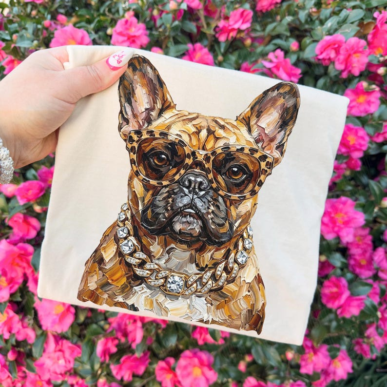 May include: A cream-colored tote bag featuring a detailed painting of a French Bulldog wearing leopard print glasses and a diamond-studded chain. The dog's fur is rendered in shades of brown and tan. The background is a vibrant display of pink flowers.