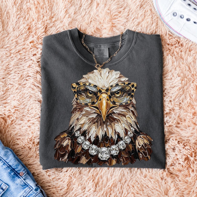 May include: Dark gray t-shirt featuring a detailed illustration of an eagle wearing leopard print glasses and a diamond necklace. The eagle's feathers are rendered in shades of brown, white, and gold. The shirt is folded on a pink, fluffy surface.