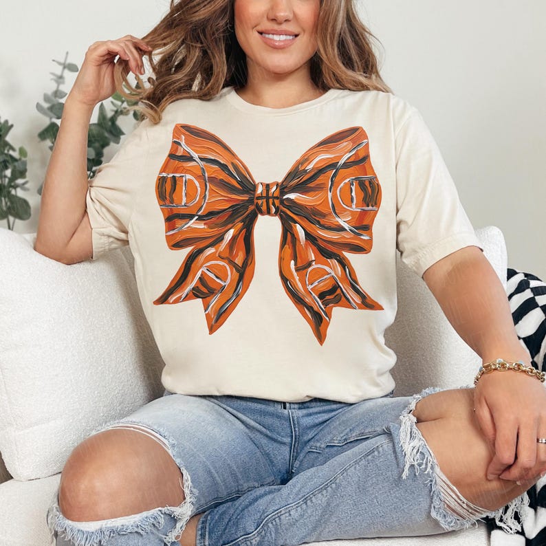 May include: Cream-colored t-shirt featuring a large bow design. The bow is orange and black, with a basketball court pattern. The shirt has short sleeves and a crew neck.