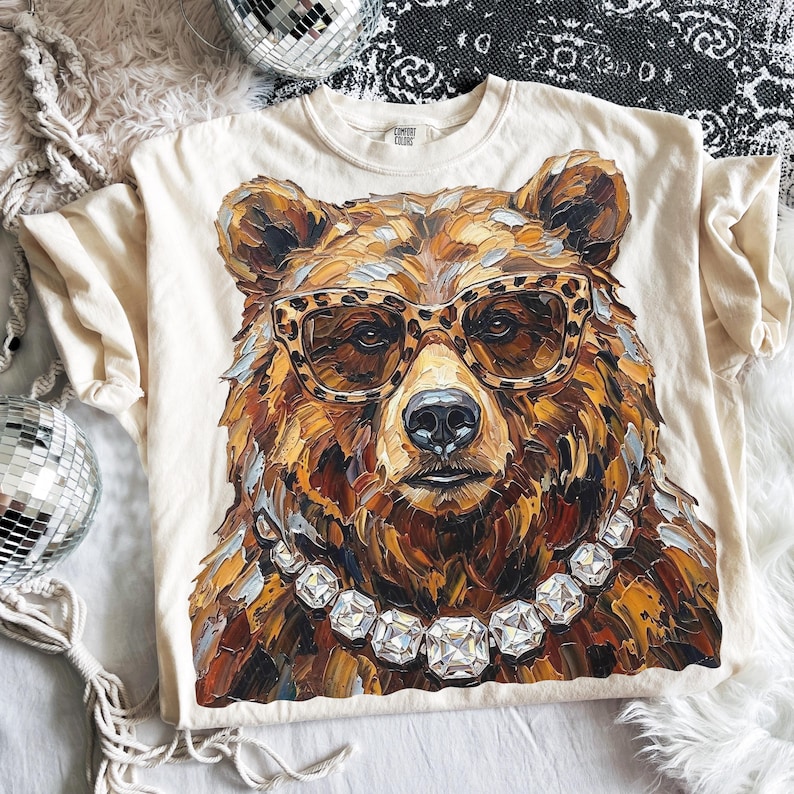 May include: Cream-colored t-shirt featuring a detailed illustration of a bear wearing leopard print glasses and a diamond necklace. The bear's fur is rendered in shades of brown, gold, and black. The shirt has a crew neck and short sleeves.