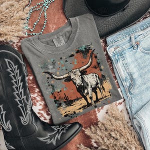 May include: A gray t-shirt featuring a longhorn illustration with a desert landscape design. The shirt is paired with blue jeans, a black cowboy hat, and a black cowboy boot. A turquoise necklace and dried grass complete the western-themed ensemble.