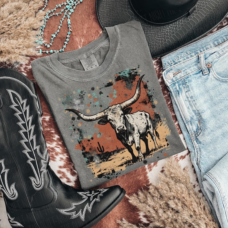 May include: A gray t-shirt featuring a longhorn illustration with a desert landscape design. The shirt is paired with blue jeans, a black cowboy hat, and a black cowboy boot. A turquoise necklace and dried grass complete the western-themed ensemble.