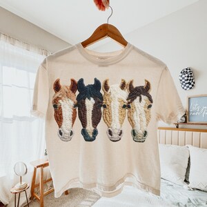 Peut inclure: Un t-shirt cr&egrave;me avec un motif brod&eacute; de quatre t&ecirc;tes de chevaux dans des tons de marron, noir et cr&egrave;me. Le t-shirt est suspendu &agrave; un cintre en bois. Le motif est centr&eacute; sur le devant du t-shirt.