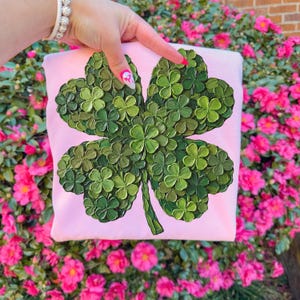 May include: A light pink sweatshirt featuring a large, textured four-leaf clover design in shades of green. The clover is the focal point, with each leaf detailed. The background is a vibrant display of pink flowers.