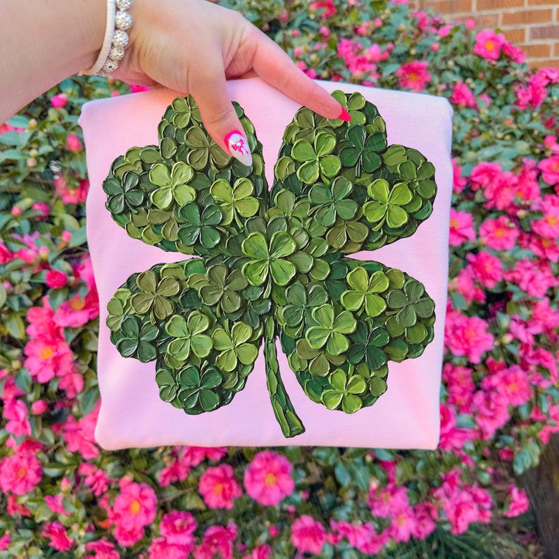 May include: A light pink sweatshirt featuring a large, textured four-leaf clover design in shades of green. The clover is the focal point, with each leaf detailed. The background is a vibrant display of pink flowers.