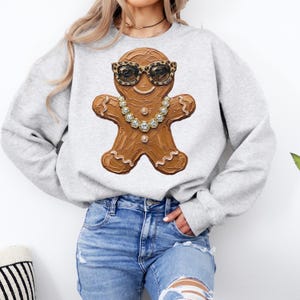 May include: A light gray crewneck sweatshirt featuring a central graphic of a brown gingerbread person wearing leopard print sunglasses and a gold-toned chain necklace with clear crystals. The gingerbread figure has white icing details.