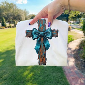 Peut inclure: T-shirt blanc avec un motif de croix peinte. La croix est marron avec des accents bleus et un n&oelig;ud bleu sarcelle. L'&oelig;uvre d'art a un aspect textur&eacute;, au coup de pinceau, cr&eacute;ant une esth&eacute;tique rustique et artistique.