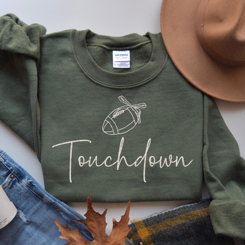 May include: An olive green sweatshirt with the word "Touchdown" in a cursive font. Above the word is a white line drawing of a football. A brown hat and blue jeans are in the background.