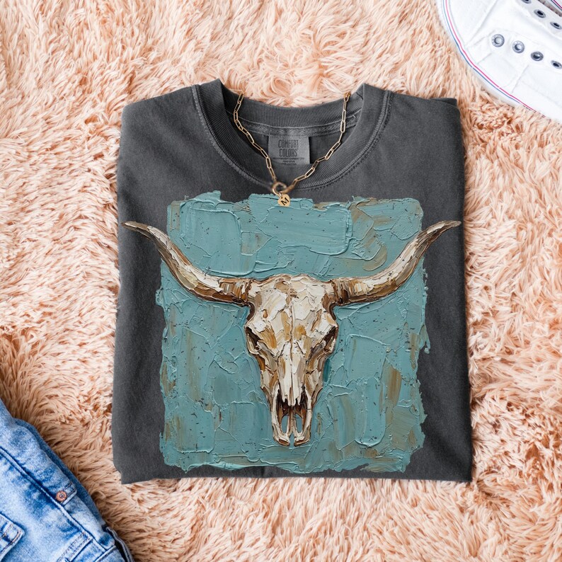 May include: Charcoal grey t-shirt with a painting of a longhorn skull on a textured teal background. The shirt has a gold chain necklace and the words "Comfort Colors" printed on the tag. Blue jeans and white trainers are also visible.