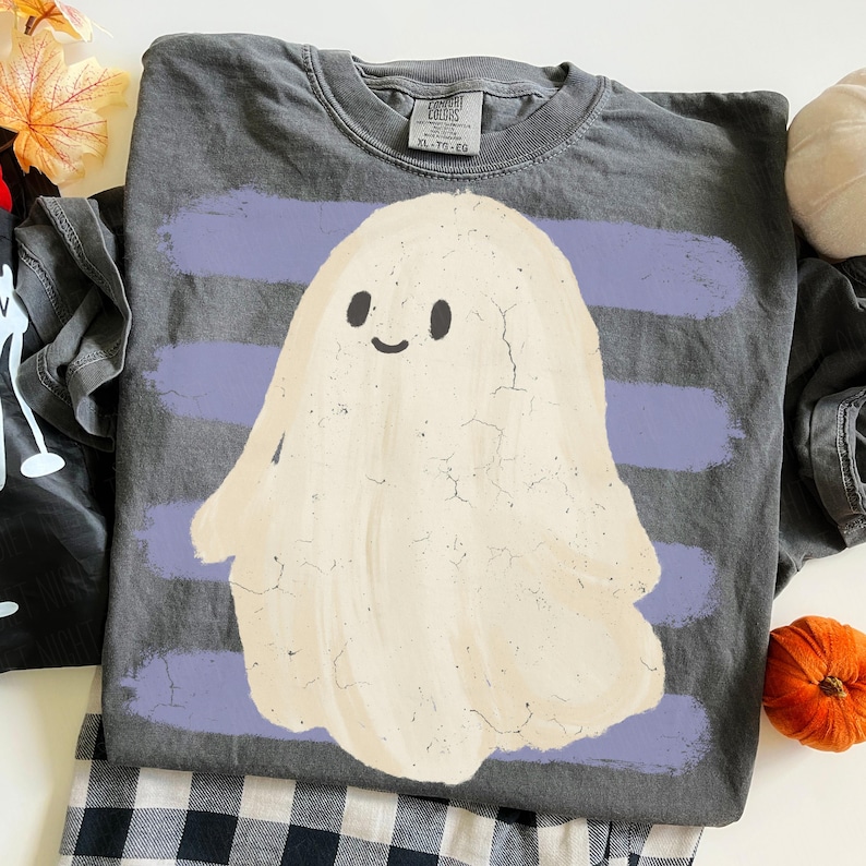 May include: A charcoal gray crew neck t-shirt featuring a large, cream-colored ghost graphic with a simple smiling face. The ghost is layered over horizontal, distressed light purple brushstroke stripes. The shirt has a tag reading Comfort Colors.
