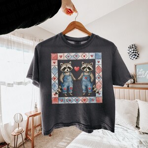 Pu&ograve; includere: T-shirt grigio scuro con una grafica di due procioni che si tengono per mano, indossando salopette blu e circondati da una cornice a forma di cuore. Il design include cuori rossi e un effetto acquerello, perfetto per San Valentino.