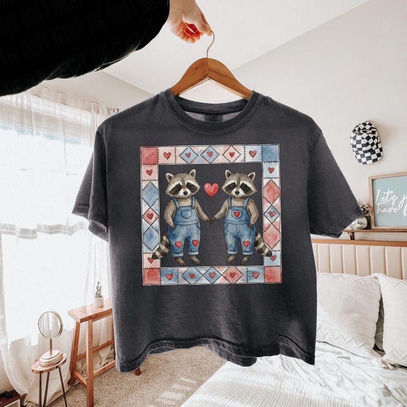 Pu&ograve; includere: T-shirt grigio scuro con una grafica di due procioni che si tengono per mano, indossando salopette blu e circondati da una cornice a forma di cuore. Il design include cuori rossi e un effetto acquerello, perfetto per San Valentino.