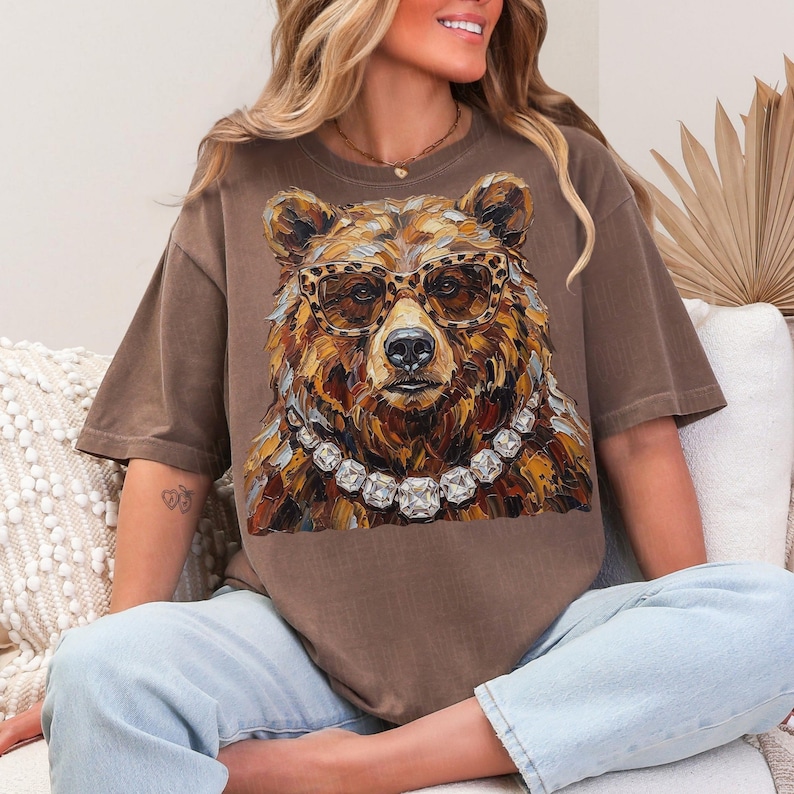 May include: Brown t-shirt featuring a detailed illustration of a bear wearing leopard print glasses and a diamond necklace. The bear's fur is rendered in shades of brown and gold. The shirt has short sleeves and a relaxed fit.