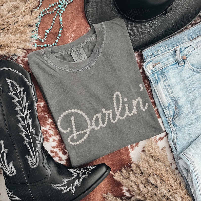 Pu&ograve; includere: Una maglietta grigia con la scritta "Darlin'" in bianco, con lettere a forma di perla. La maglietta &egrave; esposta con un paio di jeans blu, un cappello da cowboy nero e uno stivale da cowboy nero con cuciture bianche. Sono presenti anche una collana turchese ed erba secca.