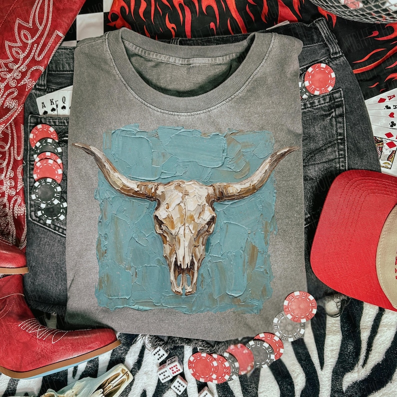 May include: Grey crew neck sweatshirt with a bull skull design on a blue textured background. The image includes red cowboy boots, a red cap, denim shorts, playing cards, and poker chips, creating a Western theme.