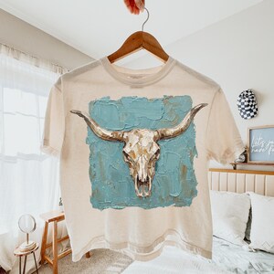 May include: A cream-coloured t-shirt displaying a detailed illustration of a bull skull with long horns. The skull is set against a textured, turquoise blue background. The shirt is hanging on a wooden hanger.