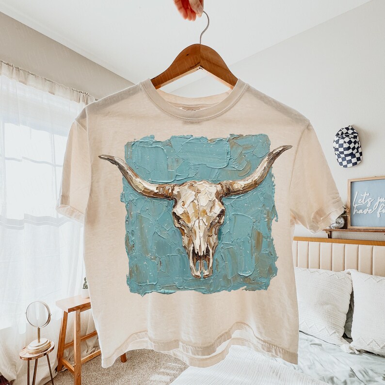 May include: A cream-coloured t-shirt displaying a detailed illustration of a bull skull with long horns. The skull is set against a textured, turquoise blue background. The shirt is hanging on a wooden hanger.