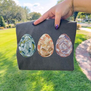 Peut inclure: Un tissu gris fonc&eacute; avec trois motifs d'&oelig;ufs de P&acirc;ques peints. Les &oelig;ufs sont dans des tons de bleu, d'orange et de violet, avec des coups de pinceau textur&eacute;s. Le tissu est pr&eacute;sent&eacute; sur un fond herbeux vert.