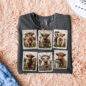 May include: Dark gray Comfort Colors t-shirt featuring six postage stamp-style images of Highland cows. Each image showcases a different cow with various accessories, set against a rural backdrop. A gold chain necklace rests on the shirt.