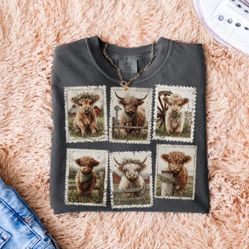 May include: Dark gray Comfort Colors t-shirt featuring six postage stamp-style images of Highland cows. Each image showcases a different cow with various accessories, set against a rural backdrop. A gold chain necklace rests on the shirt.