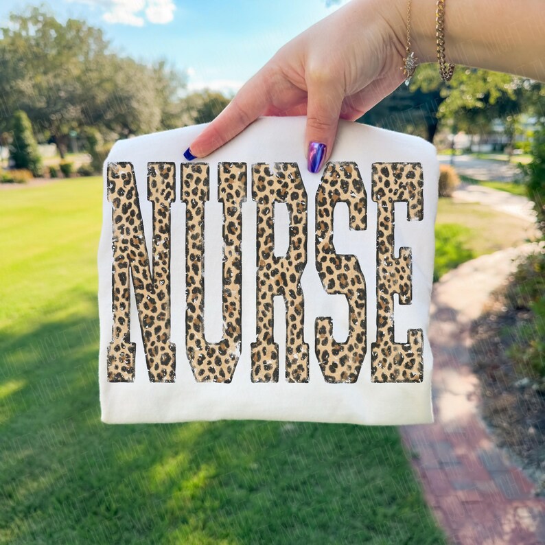 May include: A white t-shirt with the word "NURSE" printed in large, distressed leopard print letters. The shirt is held up against a background of green grass and trees.