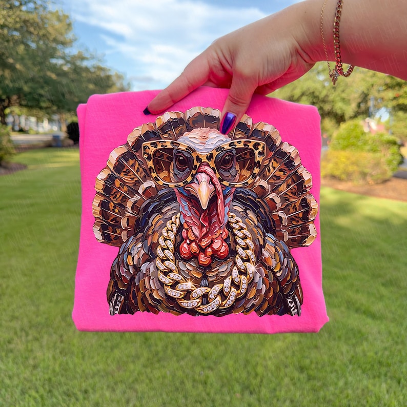 May include: A bright pink t-shirt featuring a graphic print of a turkey wearing leopard print sunglasses and a thick gold chain. The turkey has detailed brown and black feathers, a red wattle, and a beak. The design is centered on the fabric.