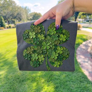 May include: A dark gray square textile with a large, textured four-leaf clover design in various shades of green. The clover is made up of many small, individual leaves, creating a 3D effect. The textile is held up against a green background.