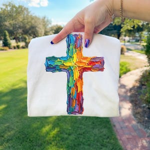 Könnte beinhalten: Weißes Sweatshirt mit einem farbenfrohen, strukturierten Kreuzdesign. Das Kreuz ist in einer Regenbogenpalette aus Rot, Orange, Gelb, Grün, Blau und Lila gehalten. Das Design hat einen dicken, pastosen Stil.