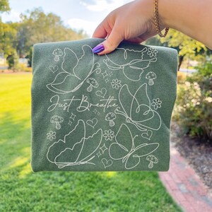 Puede incluir: Una sudadera verde con un dise&ntilde;o de l&iacute;nea blanca de mariposas, flores, corazones y setas. Las palabras "Just Breathe" est&aacute;n escritas en cursiva. La sudadera se sostiene contra un fondo de hierba verde.