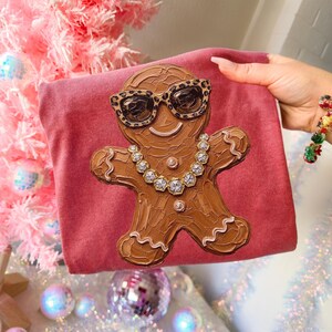 May include: A dusty rose-colored t-shirt featuring a textured, brown gingerbread person graphic. The gingerbread figure wears leopard-print sunglasses and a necklace made of clear, faceted, diamond-like stones with silver-toned settings.