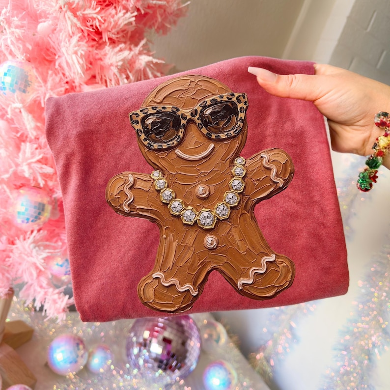 May include: A dusty rose-colored t-shirt featuring a textured, brown gingerbread person graphic. The gingerbread figure wears leopard-print sunglasses and a necklace made of clear, faceted, diamond-like stones with silver-toned settings.