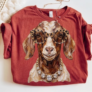 May include: A rust-colored t-shirt featuring a painterly graphic of a brown and white goat wearing leopard print glasses and a chunky gold-toned necklace with clear square crystals. The goat has a textured, brushstroke-style appearance.