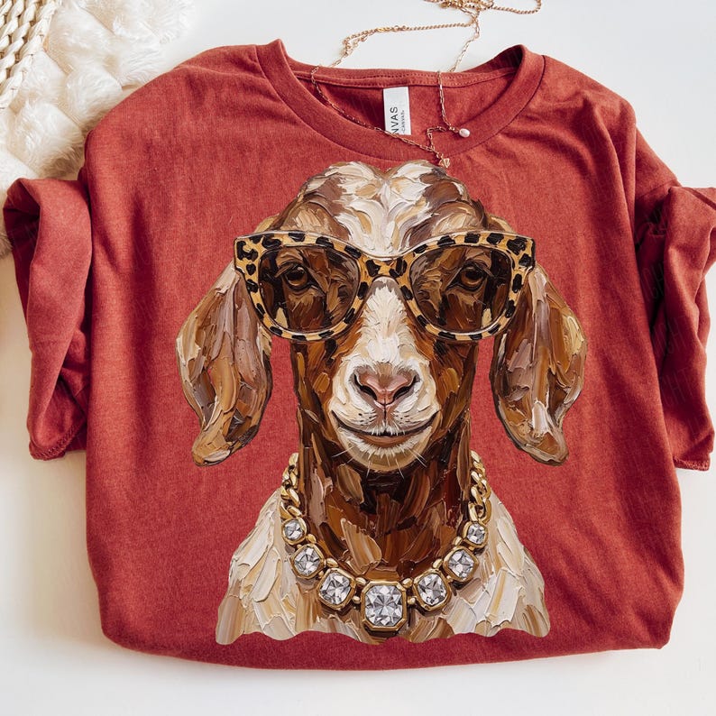 May include: A rust-colored t-shirt featuring a painterly graphic of a brown and white goat wearing leopard print glasses and a chunky gold-toned necklace with clear square crystals. The goat has a textured, brushstroke-style appearance.