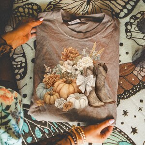 May include: A taupe-colored sweatshirt featuring a fall-themed design. The graphic includes pumpkins, flowers, and cowboy boots. A white bow is placed between the boots. The design is in shades of orange, cream, and brown.