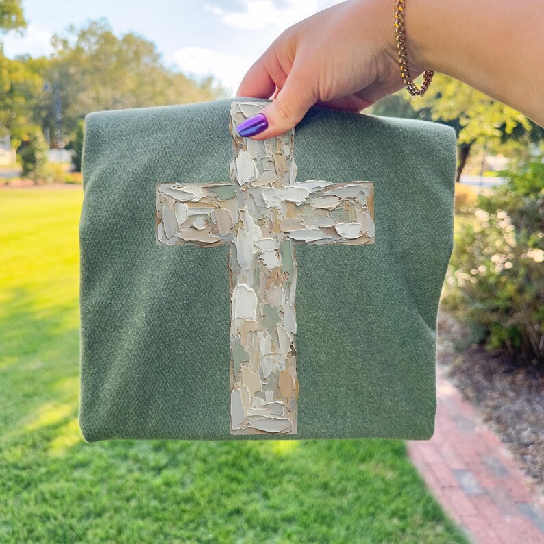 May include: A sage green square textile with a textured, painted cross design in shades of white, beige, and brown. The cross is the focal point, with visible brushstrokes. The textile is held up against a blurred outdoor background.