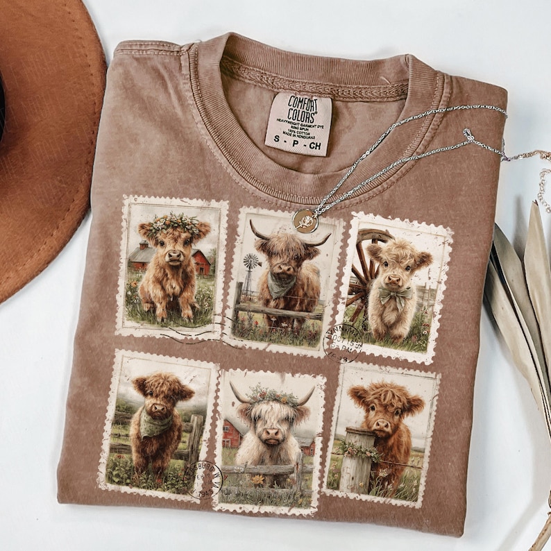 May include: A brown Comfort Colors t-shirt featuring a collage of six postage stamp-style images of Highland cows. The cows are depicted in various poses, some wearing floral crowns or bandanas. A silver necklace with a pendant rests on the shirt.