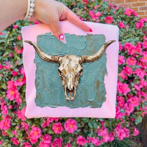 May include: A light pink sweatshirt with a gold-coloured steer skull and long horns. The skull is set against a textured teal background. The sweatshirt is held up in front of a backdrop of pink flowers.